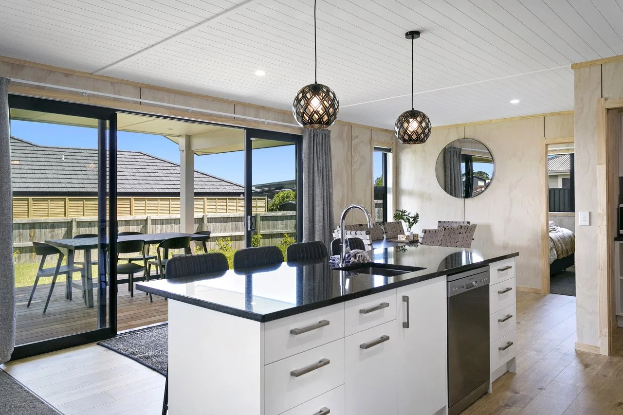 Modern kitchen with an island, black countertop, white cabinets, stainless steel dishwasher, and two pendant lights. Adjacent dining area with seating and a sliding glass door leading to an outdoor patio with dining table and chairs. Bedroom visible 