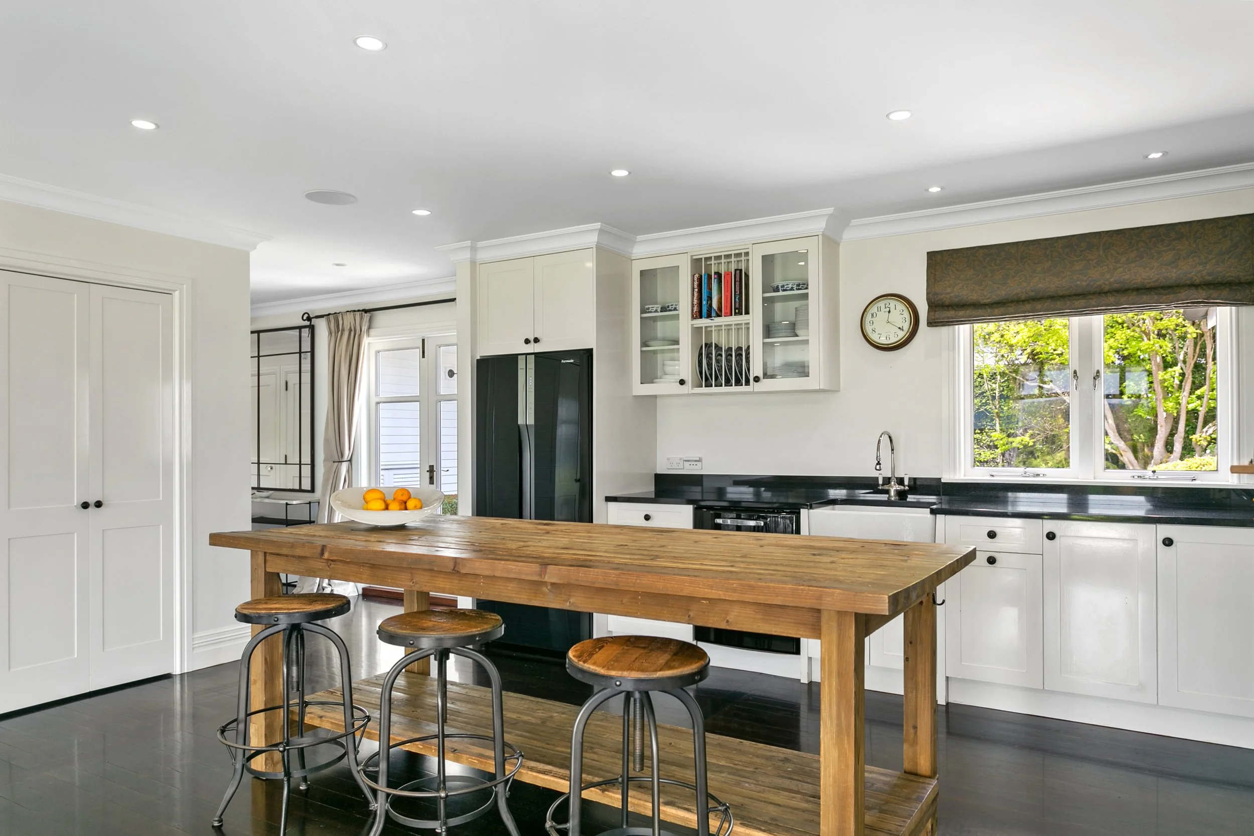 Kitchen with white cabinets, black countertops, a wooden island with three bar stools, black refrigerator, open shelves with dishes and cookbooks, window with green trees outside, and a wall clock.