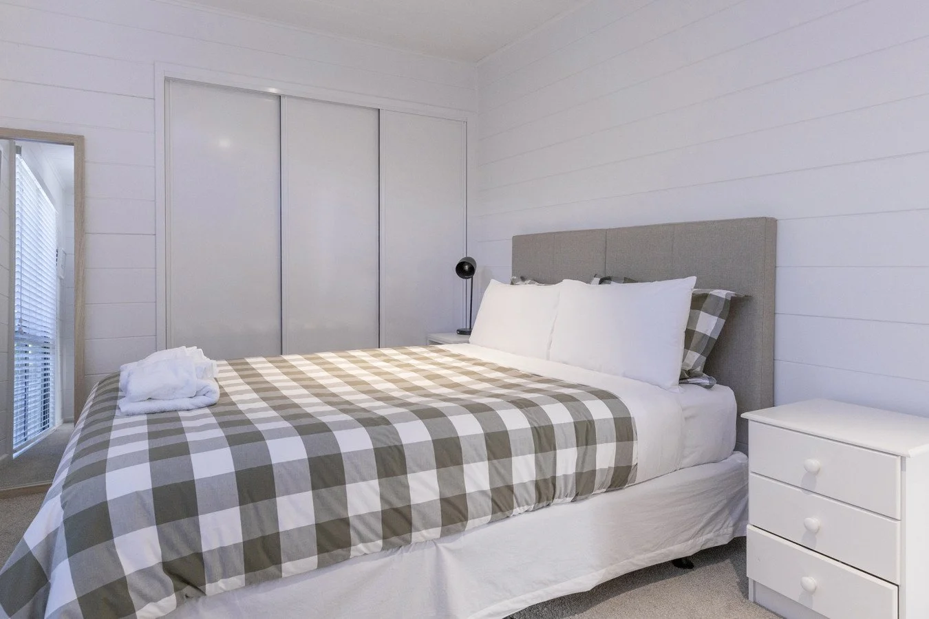 A neatly made bed with a gray checkered duvet, three large white pillows, and an gray upholstered headboard in a minimalist white bedroom with a white nightstand and black bedside lamp.