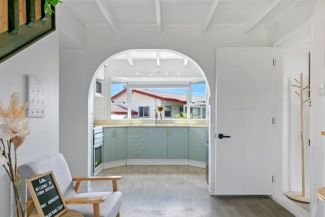 View of a cozy kitchen seen through an arched doorway, with pastel green cabinets, a window with a view of neighboring houses, and a flower vase on the counter.