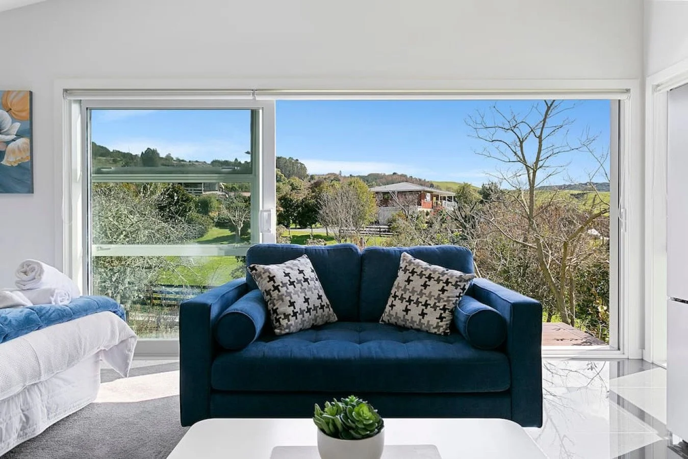 Living room with large window overlooking a green outdoor landscape, including trees and hills, with a blue sofa and patterned cushions.