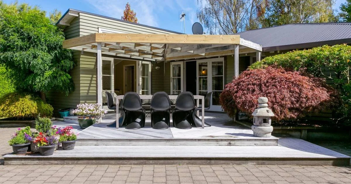 Backyard patio with outdoor dining table, black chairs, potted flowers, a Japanese stone lantern, and surrounding green shrubs and trees.