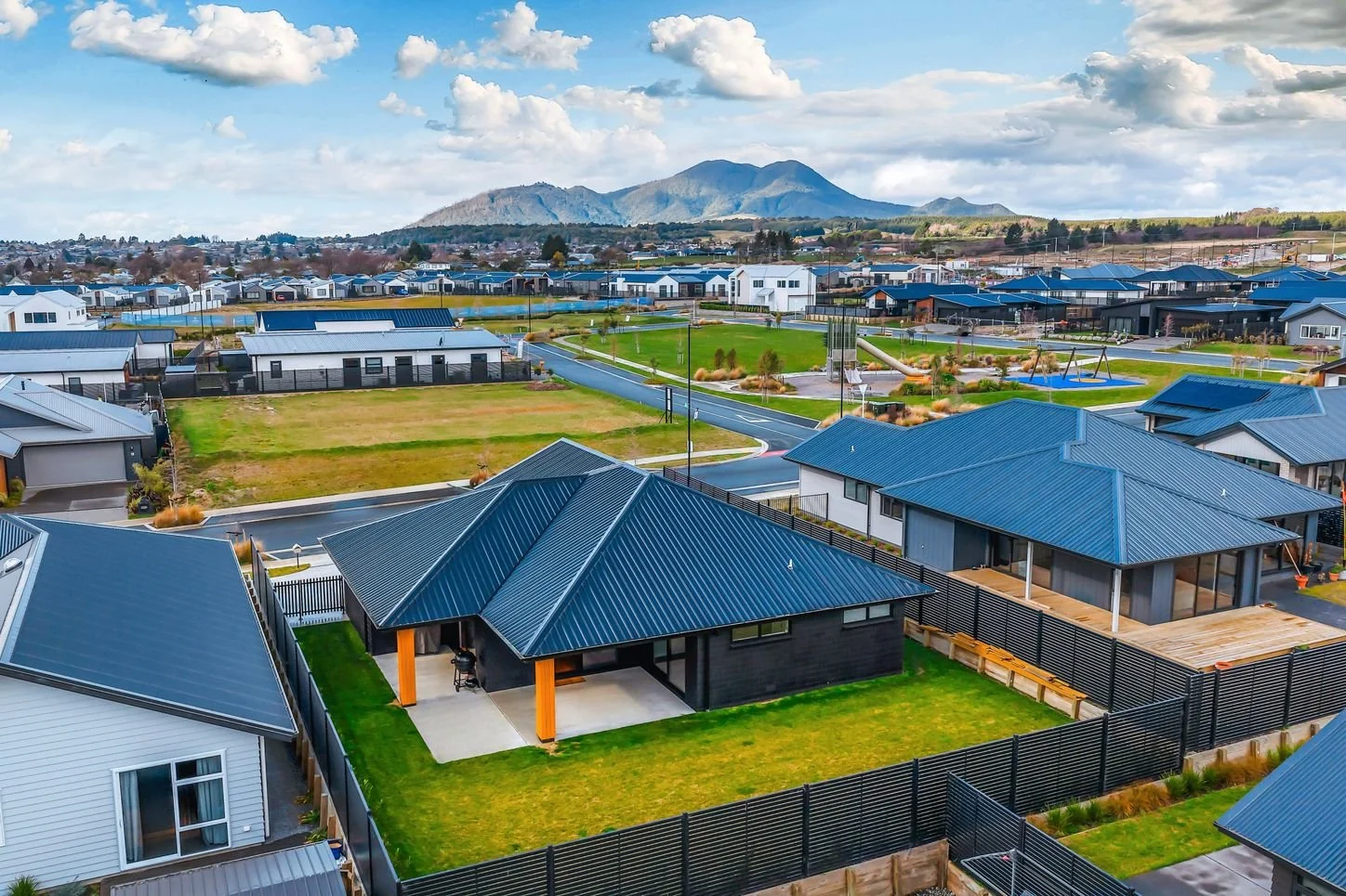 Aerial view of a modern suburban neighborhood with houses, streets, grassy areas, a playground, and mountains in the background.