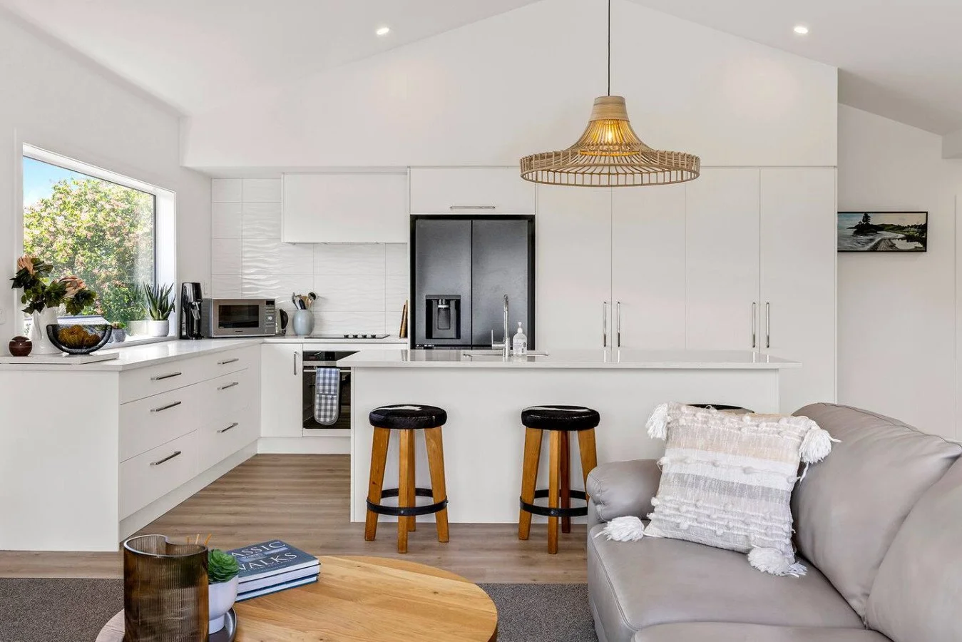 Modern white kitchen with bar stools, black refrigerator, and a gray sofa with a decorative pillow, in an open living space with a window and artwork on the wall.