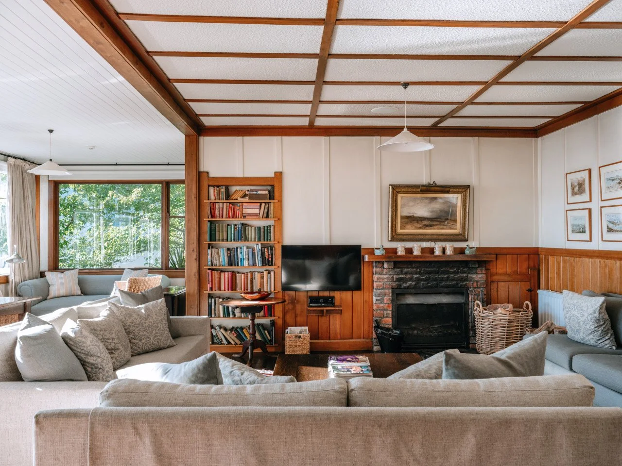 Living room with beige sofas, a brick fireplace, a mounted flat-screen TV, wooden bookshelves, a window with curtains, framed art on the walls, and a ceiling with wooden beams.