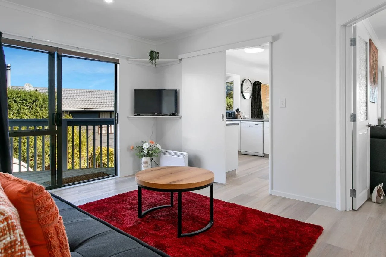 Living room with sliding glass door opening to balcony, black sofa with orange cushion, round wooden coffee table on red rug, small flat-screen TV on white wall shelf, view of kitchen through an open doorway, and a door leading to another room.