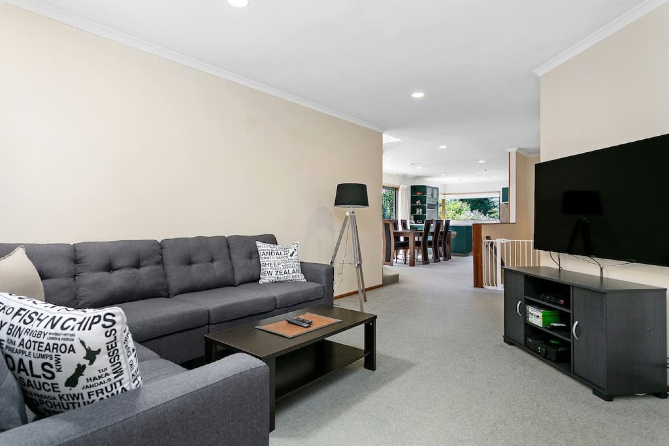Living room with a gray sofa, a black coffee table, and a TV on a black stand. In the background, a dining area with a wooden table and chairs is visible. The room has beige walls and carpeted floor.