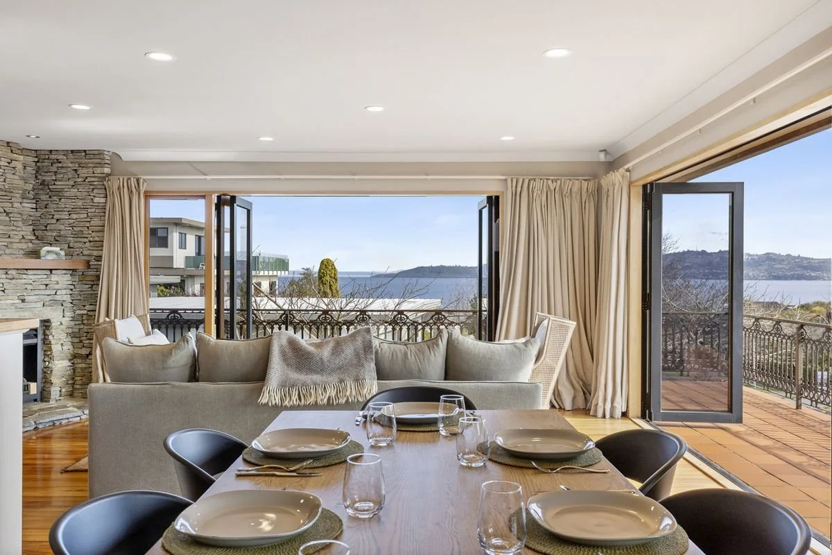 Living room with large windows revealing a balcony and water view, dining table set with plates and glasses, beige sofa with blanket, stone fireplace, and neutral curtains.