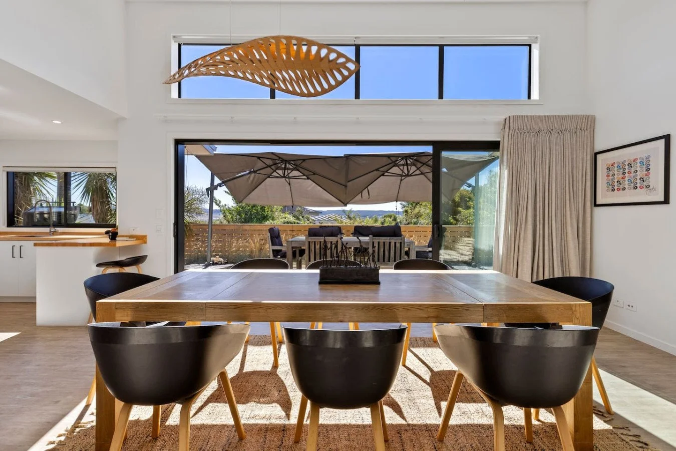 Modern dining room with wooden table, six black chairs, and large sliding glass doors leading to an outdoor patio with umbrella and seating, bright natural light, and minimalist decor.