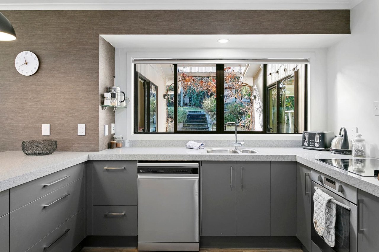 Modern kitchen with gray cabinets, a large window overlooking a backyard with steps and trees, and various small appliances on the countertop.