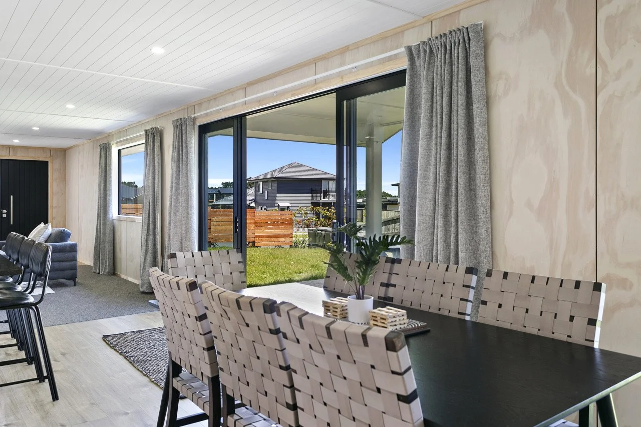 Dining area with a black table, woven chairs, a potted plant, and view of an outdoor yard through sliding glass doors.
