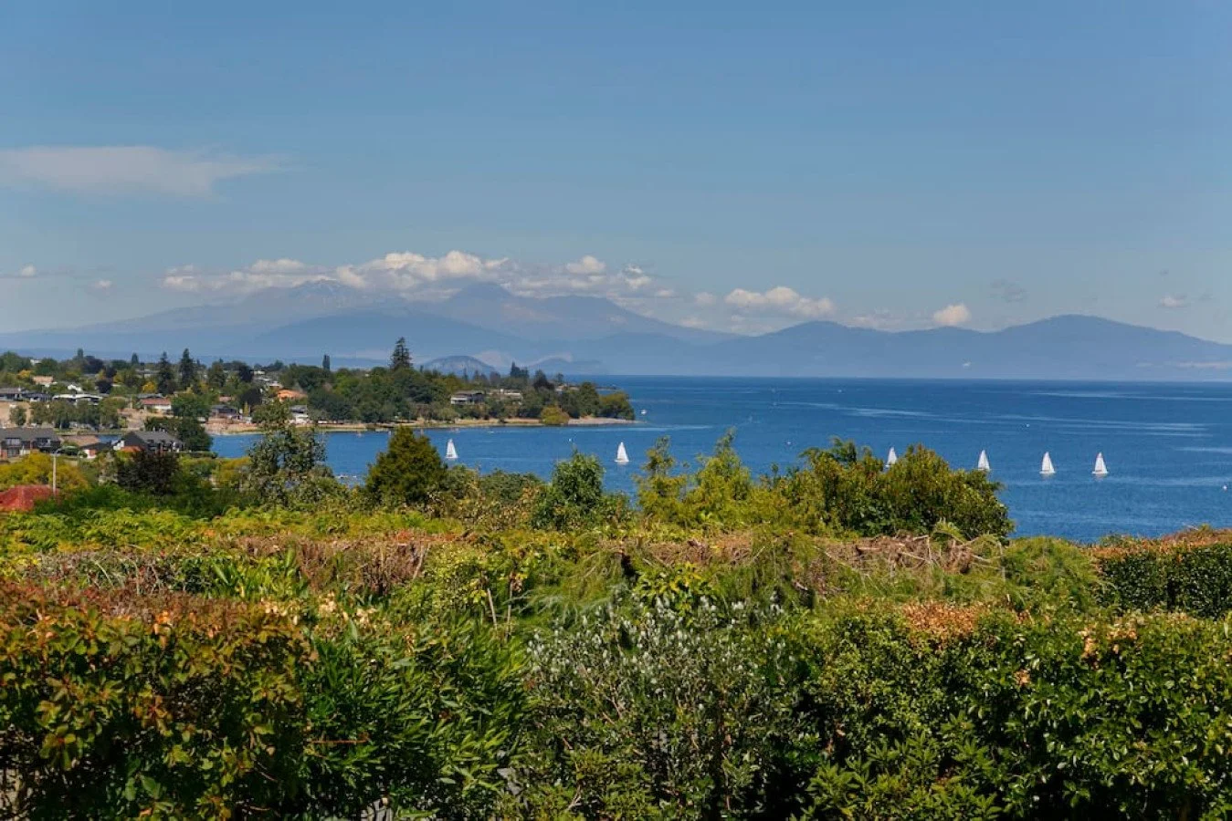 Scenic view of a coastal landscape with a body of water, sailboats, green vegetation, and a distant mountain range under a partly cloudy sky.