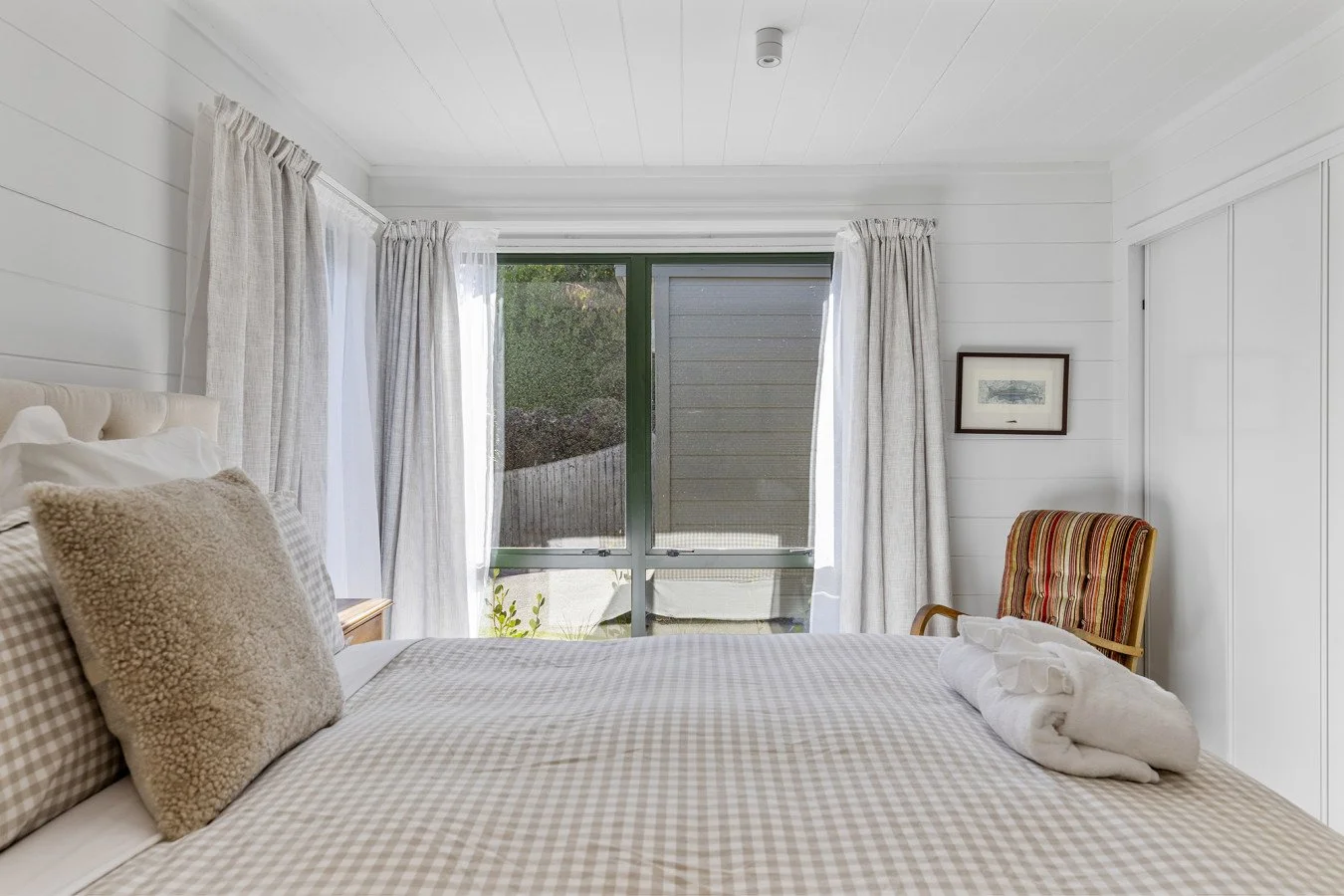 A cozy bedroom with a bed, beige checkered bedding and pillows, white curtains, and a window with greenery outside. There's a framed picture on the wall and a striped armchair in the corner.