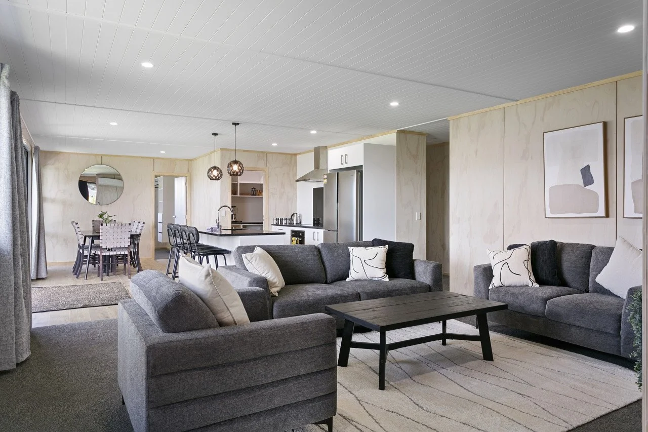 Spacious open-concept living room and dining area with grey sofas, white pillows, black coffee table, and abstract wall art, connected to a kitchen with white cabinets, black countertops, and a dining table.