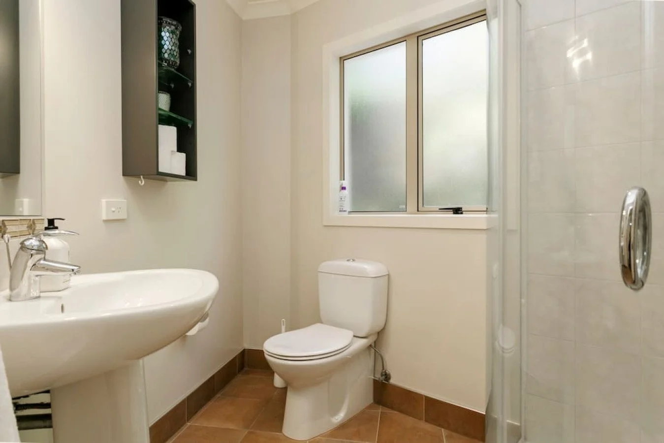 Bathroom with white toilet, sink, black wall cabinet, frosted window, beige tiled floor, and a glass shower door.