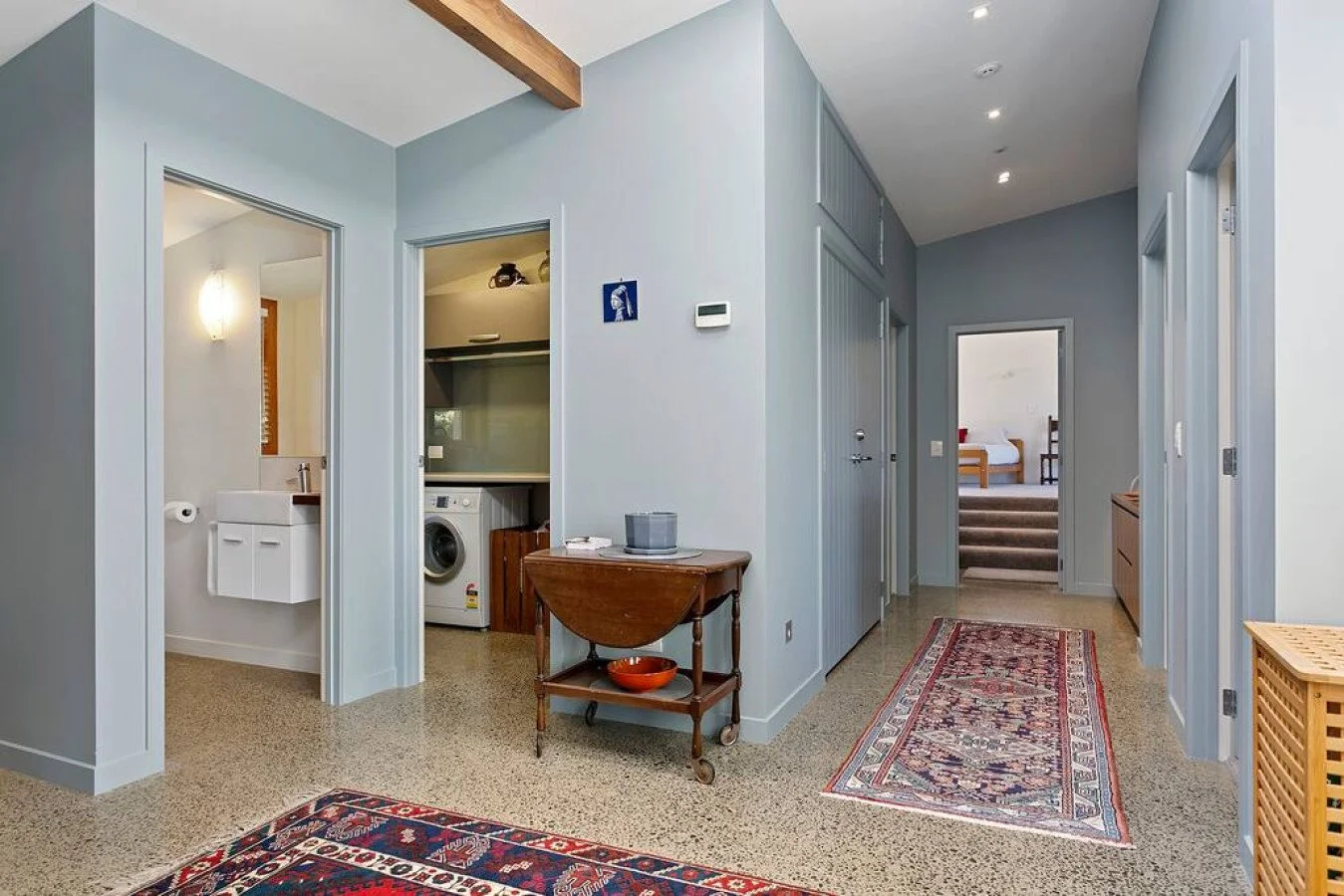 Interior view of a house hallway with light blue walls, two patterned rugs, a wooden side table, and doorways leading to laundry area, bathroom, and bedrooms.