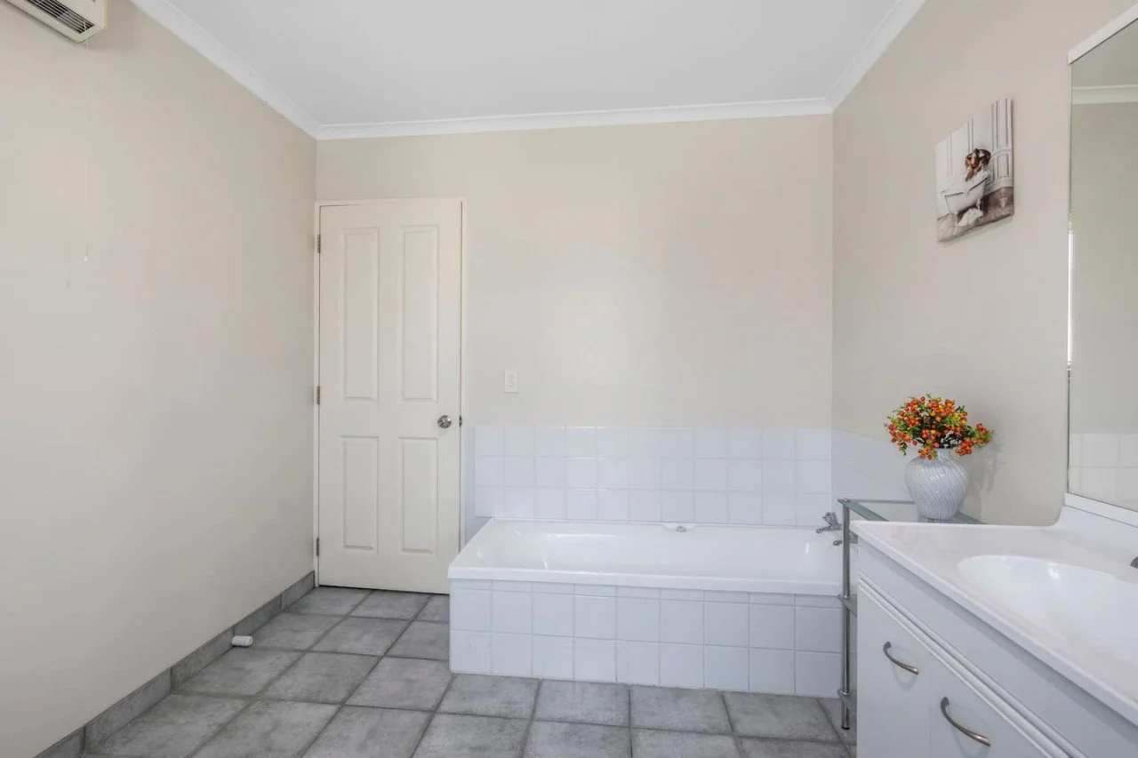 Bathroom with a closed door, a white bathtub, a sink with a white cabinet, a large mirror, a vase with flowers, and wall art with a woman in a bathtub.