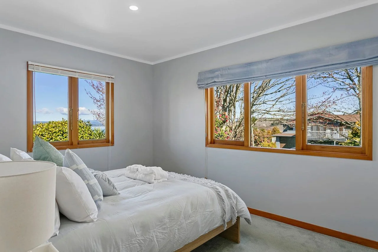 A bedroom with light gray walls, a bed with white bedding and pillows, and three large windows showing trees, a house, and a clear blue sky outside.