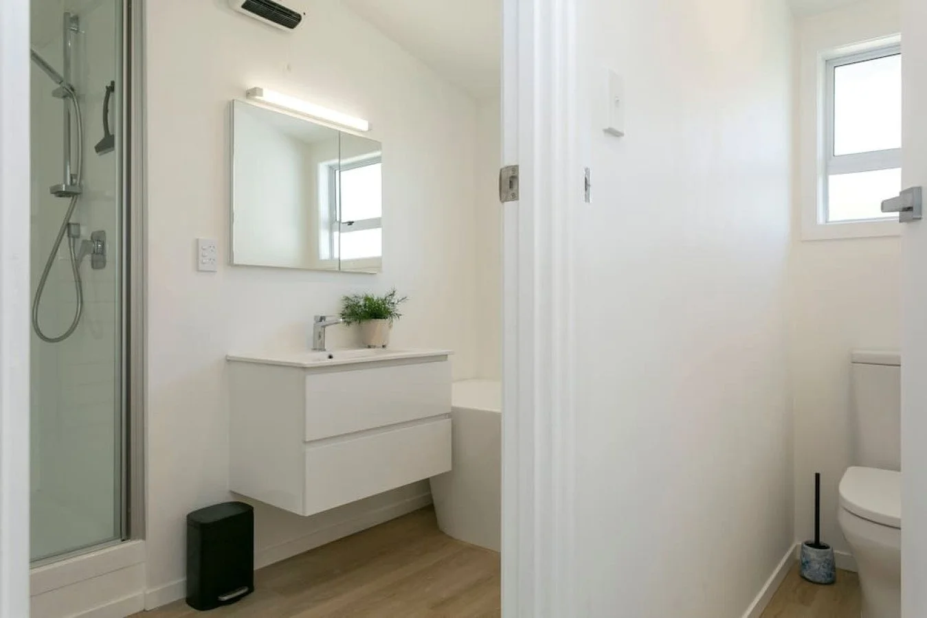 A small modern bathroom with a white vanity, a mirror, a shower stall, a toilet, a window, and light wood flooring.