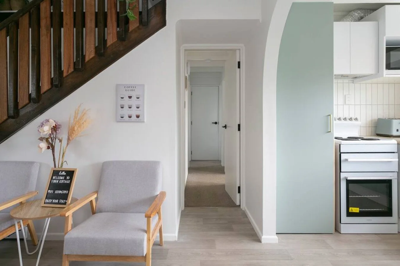 Interior view of a cozy home with a small sitting area, white walls, and a kitchen with white cabinets, a stove, and a microwave. A staircase with dark wood railing is in the corner, and there is a welcome sign and decorative flowers on a side table.