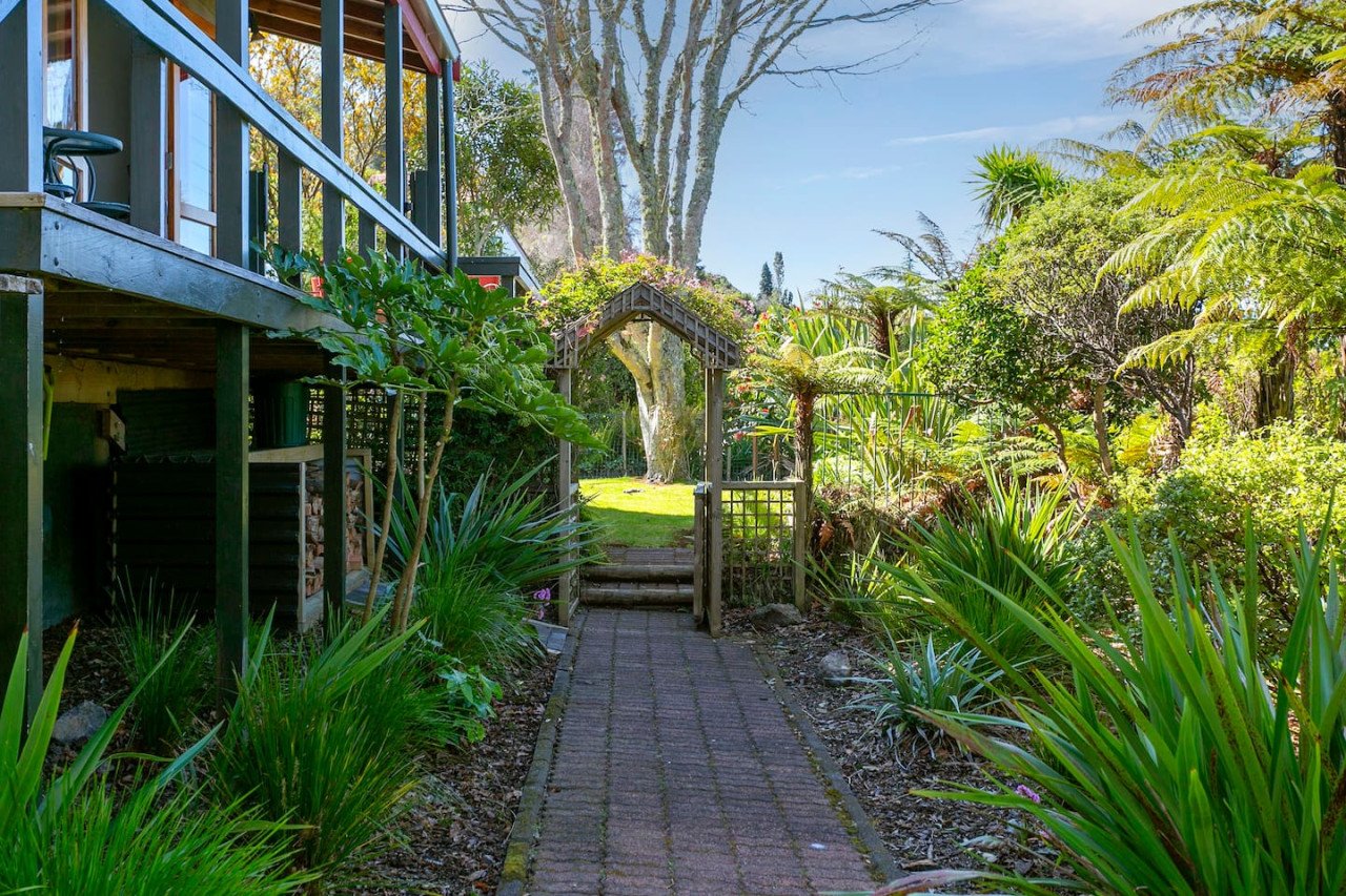 A garden with a brick pathway leading to a wooden gate and a lush green lawn surrounded by various plants and trees, with a house partially visible on the left.