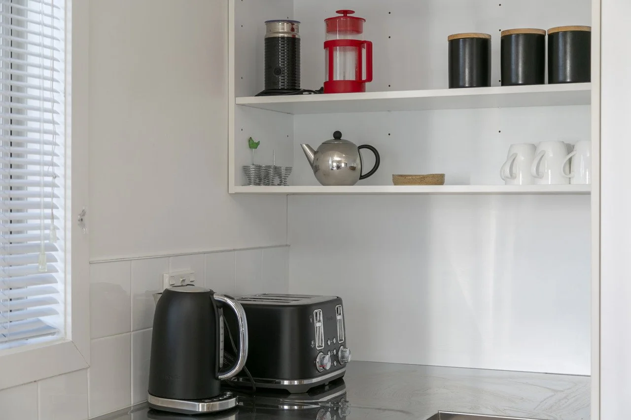 Kitchen corner with a silver electric kettle and black toaster on a gray countertop, white cabinets, and open shelves with pots, mugs, a teapot, and kitchen appliances.