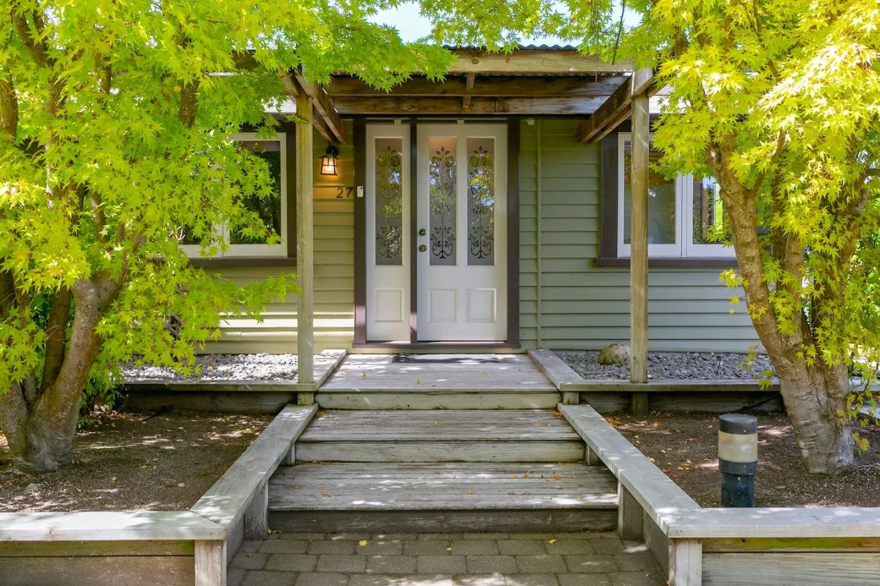Front porch of a house with a double door entrance, surrounded by green trees and wooden steps leading up to the door.