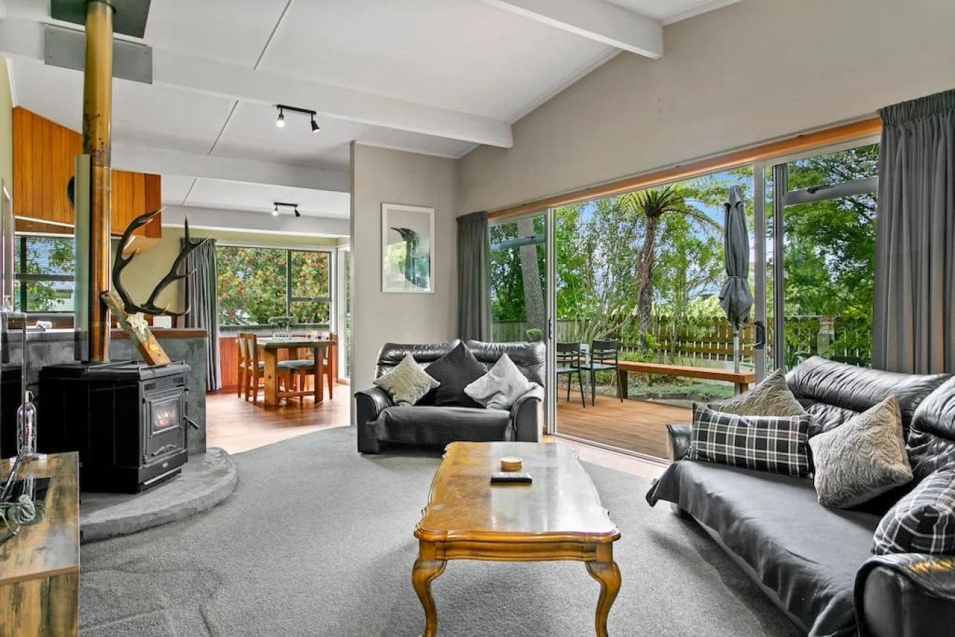 Living room with black leather couches, a wooden coffee table, and large sliding glass doors leading to a wooden deck with outdoor seating, surrounded by green trees.