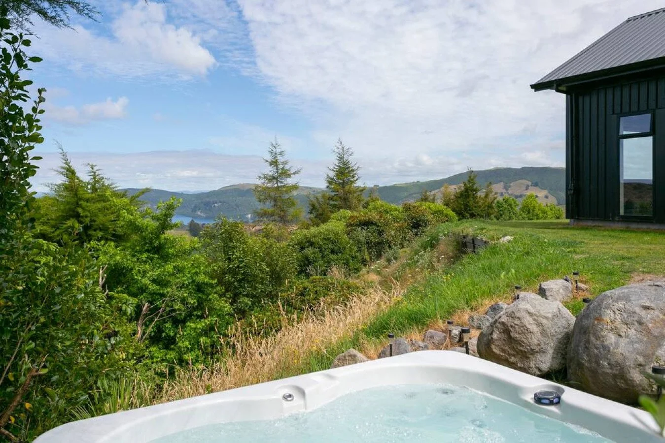 Outdoor hot tub on a deck overlooking lush green trees, rolling hills, and a lake with a partly cloudy sky.