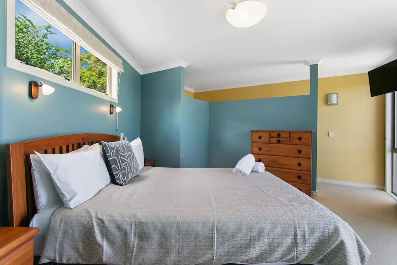 A bedroom with a bed, pillows, and a rolled towel on top. The room has light blue, yellow, and beige walls, a wooden dresser, and windows with trees outside.