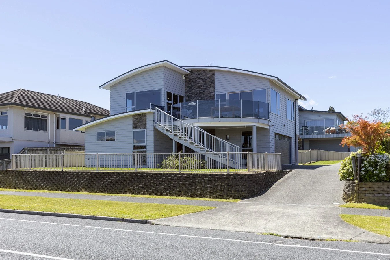 Modern multi-story house with large windows, white siding, stone accents, a balcony with glass railings, and a driveway leading to a garage, surrounded by a fenced yard and neighboring homes.