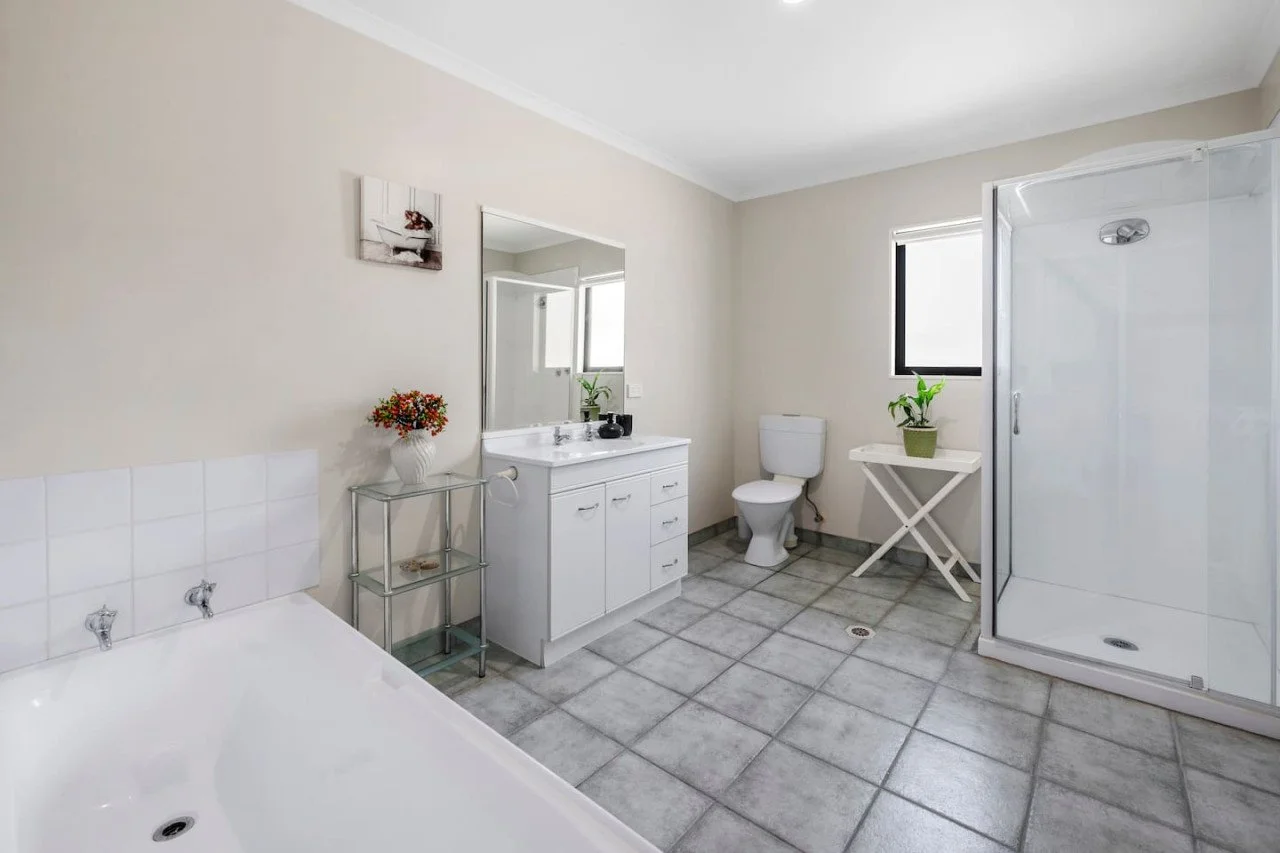 Bathroom with a bathtub, sink with a mirror, toilet, shower stall, small side table with a potted plant, and tiled flooring.