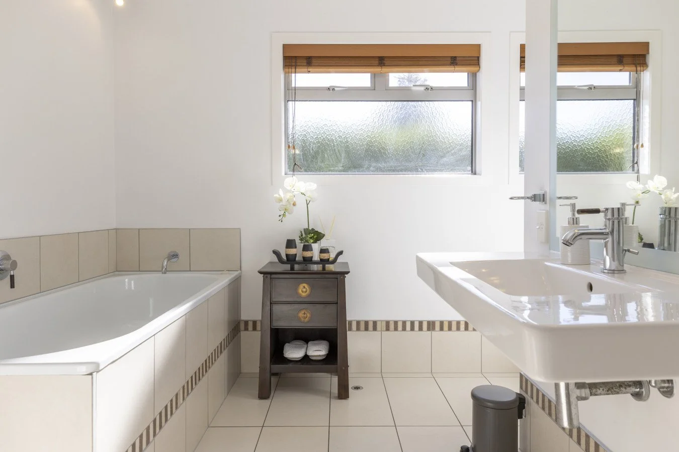 Bathroom with a bathtub on the left, a sink with a mirror on the right, a small dark wood table with flowers in the center, and frosted windows above.