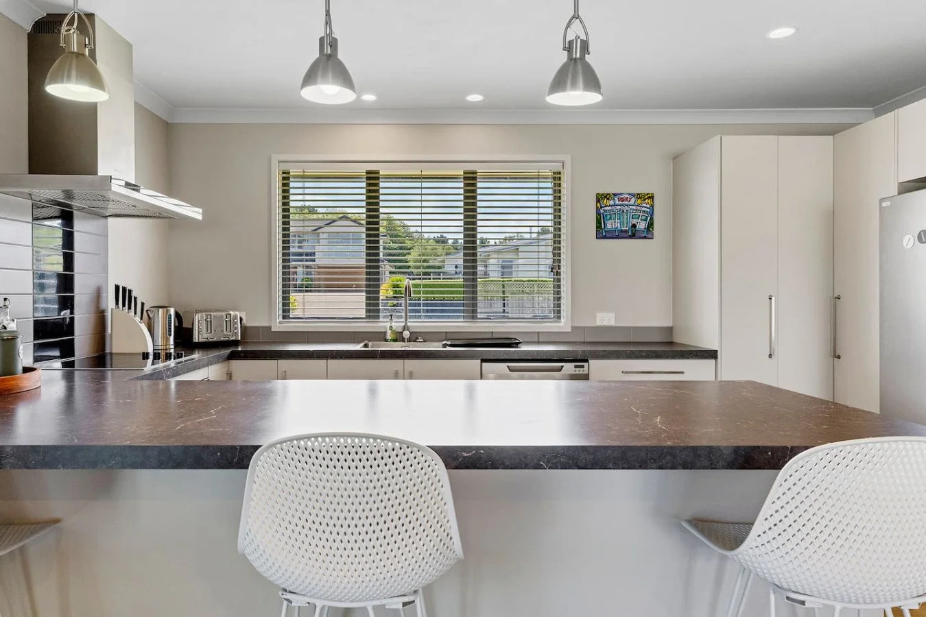 Modern kitchen with white cabinets, a window with blinds, a black countertop, and pendant lights, with a breakfast bar and white perforated chairs.