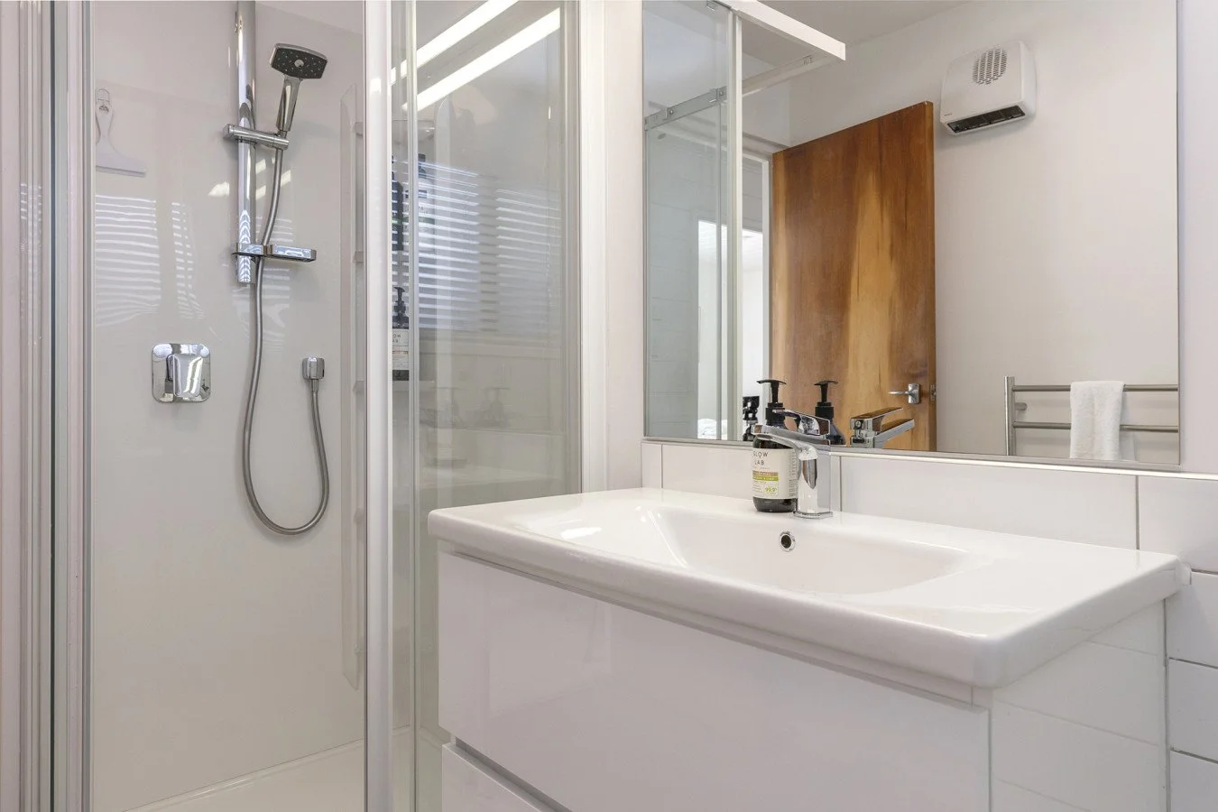 Modern bathroom with white sink, large mirror, wooden door, shower area with glass enclosure, wall-mounted heater, and towel rack
