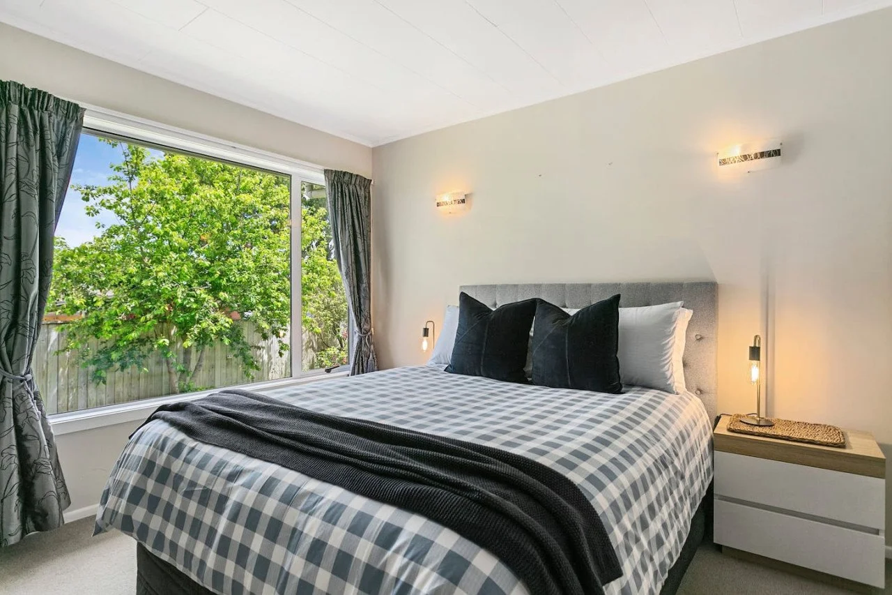 A bedroom with a large window showing green trees outside. The bed has a checkered bedspread, three black pillows, and two white pillows. There is a nightstand with a lamp beside the bed.