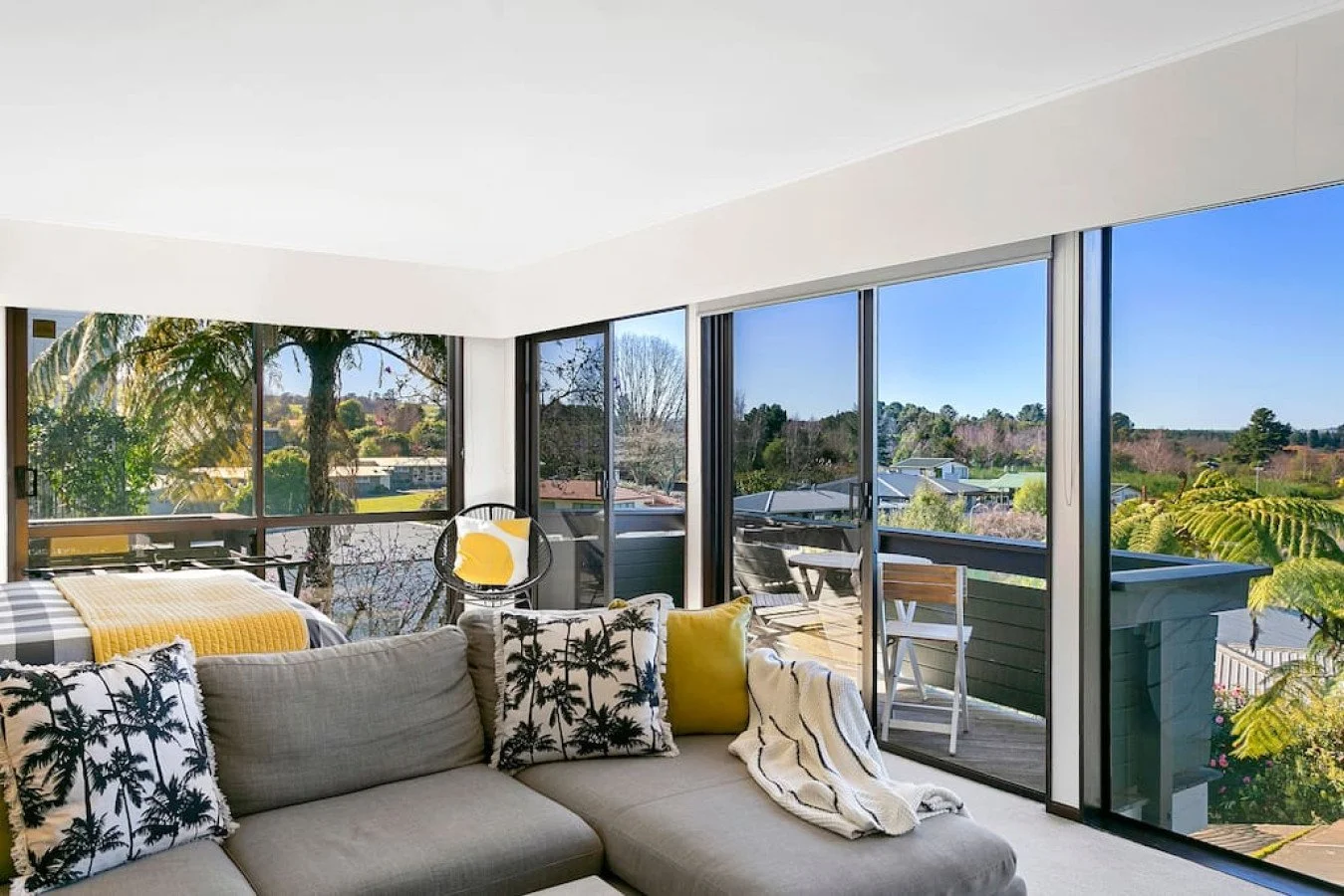 Living room with large glass sliding doors opening to balcony, view of trees and neighborhood, beige sofa with black and white pillows, yellow accent pillows, and throw blanket.