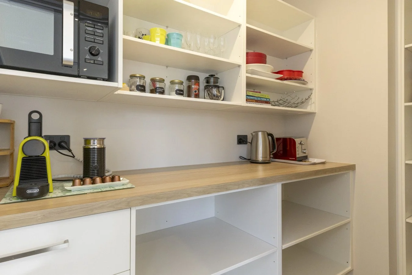 Kitchen counter with small appliances, open shelves with dishes, mugs, jars, and glasses, a microwave on the top left, a yellow espresso maker, a jar, an electric kettle, and a red toaster on the countertop, and empty open shelves below.