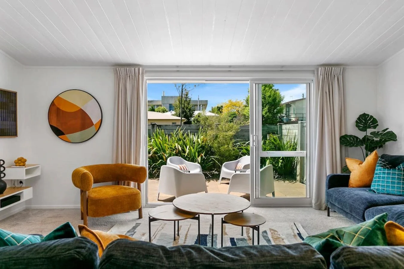 Living room with sliding glass door leading to a patio with outdoor chairs, view of greenery and neighboring houses, decorated with contemporary furniture and artwork.