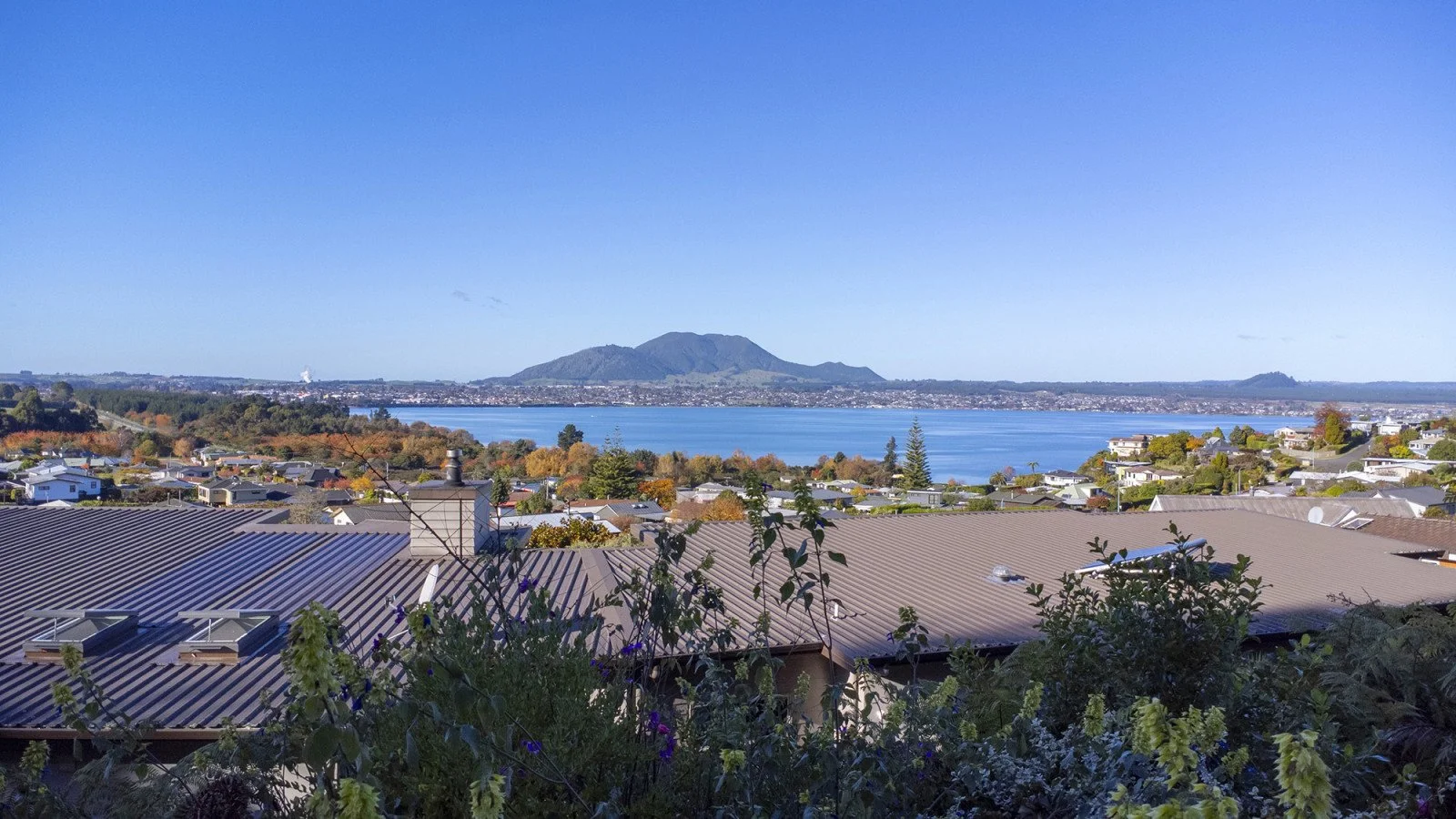 A scenic view of a lake with a mountain in the background, houses and trees in the foreground, and a clear blue sky.