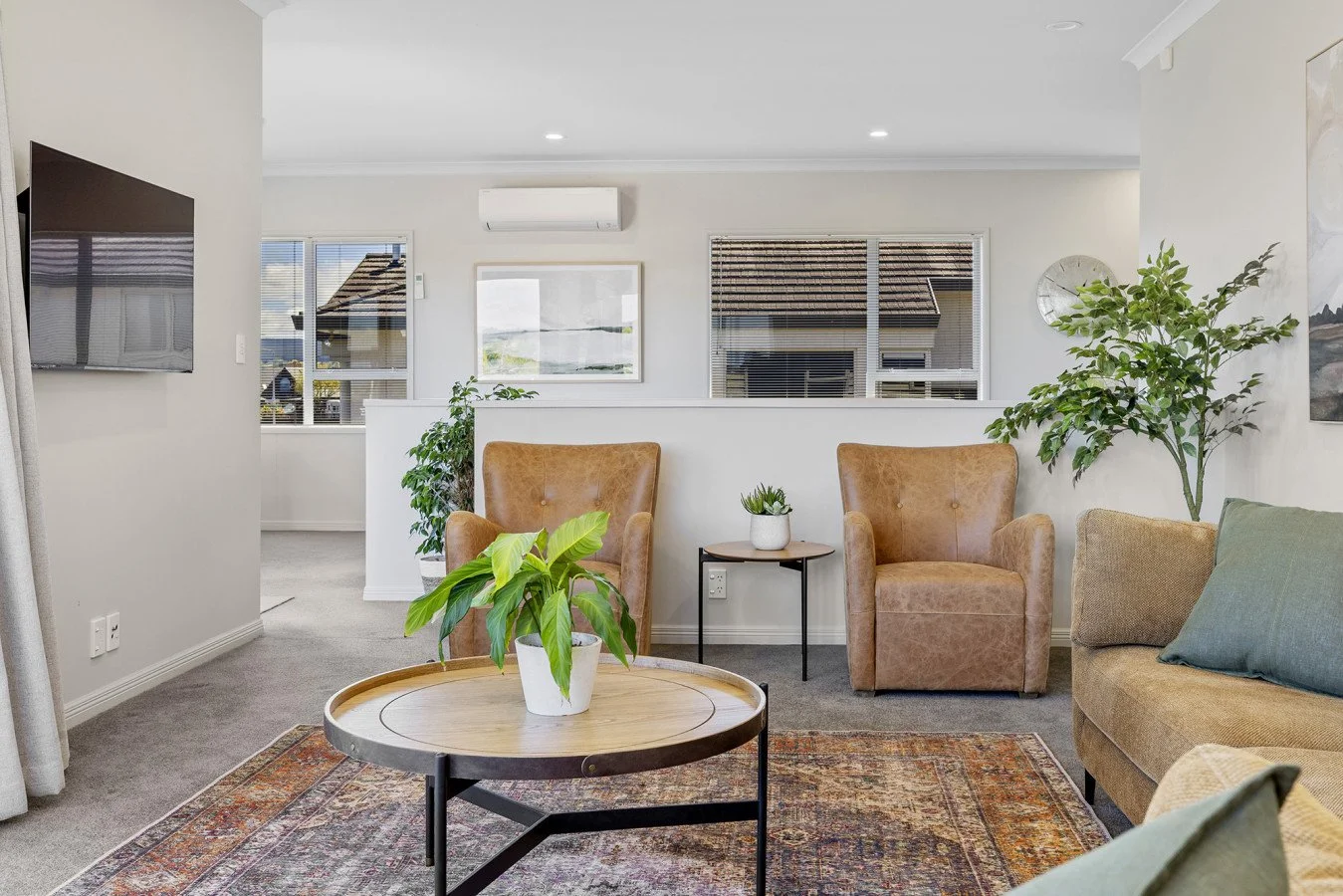 Living room with beige armchairs, a green sofa, potted plants, a round coffee table with a potted plant, window views, and wall art.