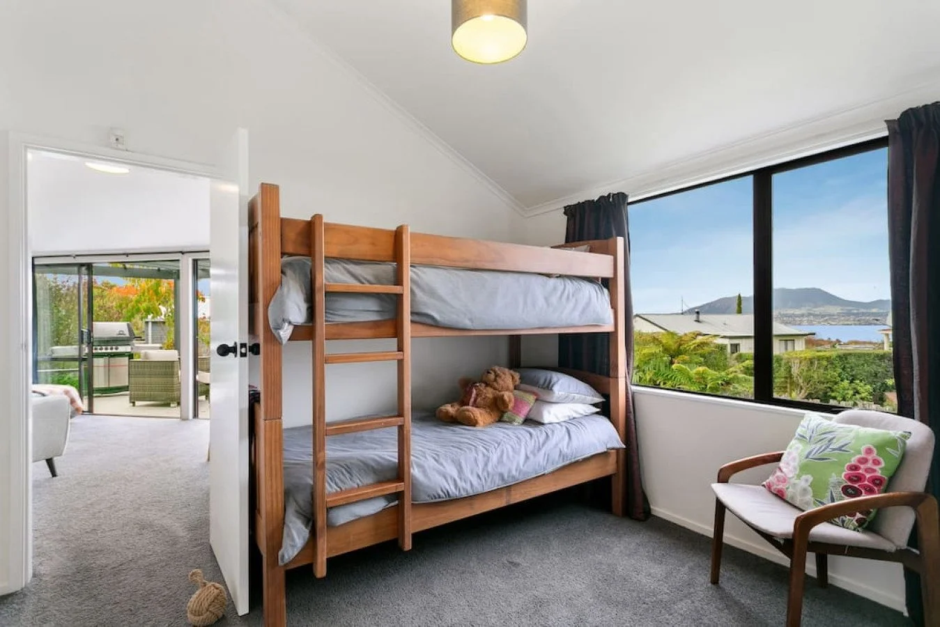 Bedroom with wooden bunk bed, teddy bear and pillow on lower bed, large window with a view of greenery, houses, mountains, and water, armchair with floral pillow, ceiling light, and an adjacent room with outdoor patio.