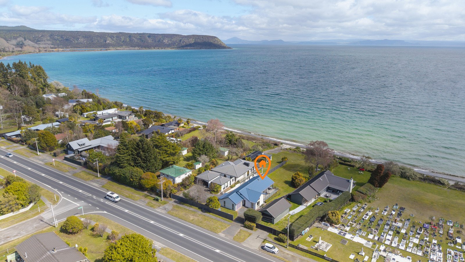Aerial view of a coastal neighborhood with houses, trees, a road, and a cemetery, overlooking the ocean with hills in the background.