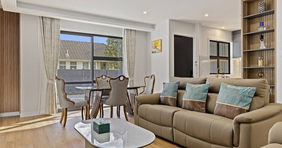 A modern living room with a beige sofa, decorative pillows, a white coffee table, a dining table with six chairs, large windows with curtains, and built-in shelves on the wall.