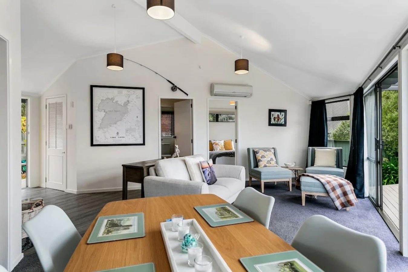 Living room with white walls, a ceiling with ceiling lights, a closet, and sliding glass doors leading to a balcony with outdoor greenery. There are multiple chairs and a sofa with cushions, a wooden table with placemats and glasses, and framed art o