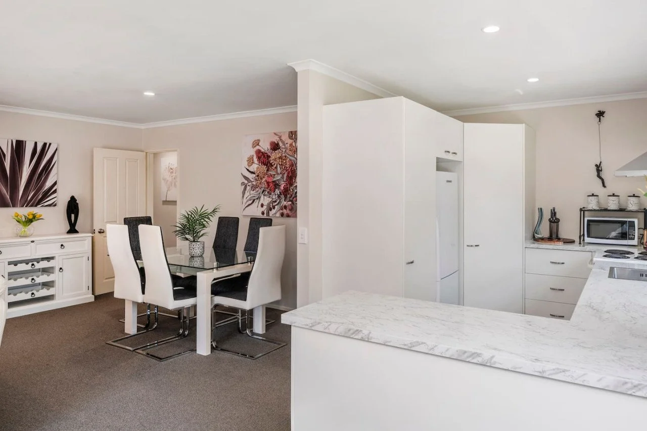 Interior of a modern, open-plan kitchen and dining area with white cabinetry, marble countertops, a dining table with six chairs, and decorative artwork on the walls.
