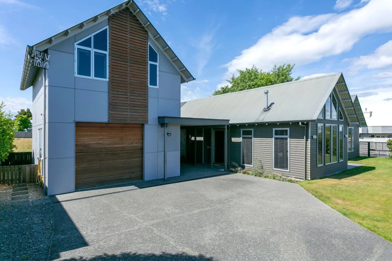 Modern house with a mix of wood and siding exterior, a steep gable roof, large windows, and a paved driveway under a blue sky with clouds.