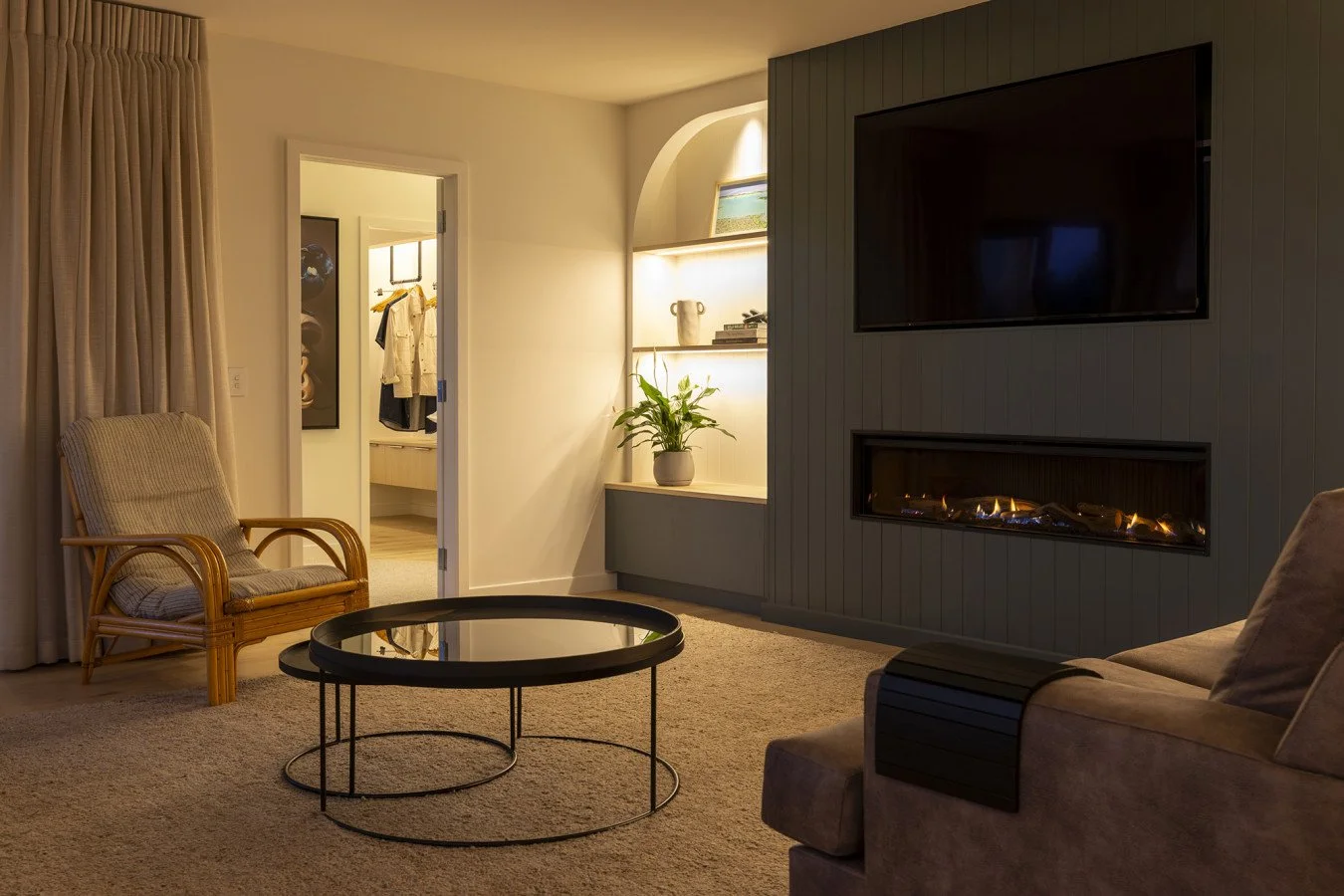 Living room with beige carpet, cushioned armchair, beige sofa with black accent, black round coffee table, flat-screen TV, modern fireplace, built-in shelves with plant and decorative items, doorway leading to bedroom with clothes, curtains on the wi