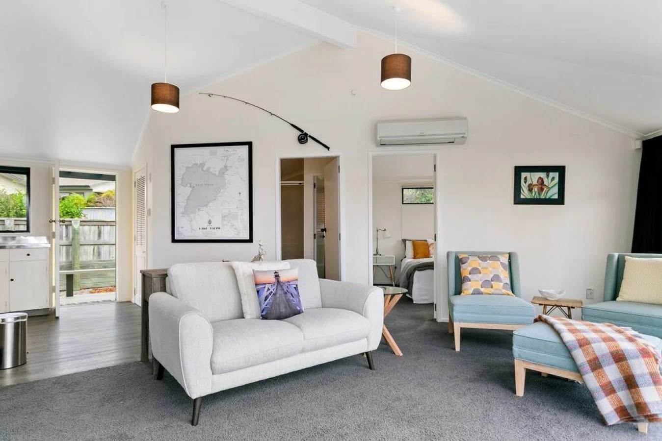 Living room with white couch, two light blue armchairs, and a dark gray carpet. Artworks on the walls, one of a world map and one of flowers. Open door leads outside, and a close door reveals another room with a bed. Various pendant lights hang from 