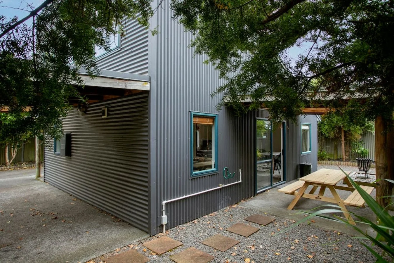 A modern metal building with corrugated steel siding, surrounded by trees and a fenced backyard. There is a sliding glass door, a window, and a wooden picnic table outside.